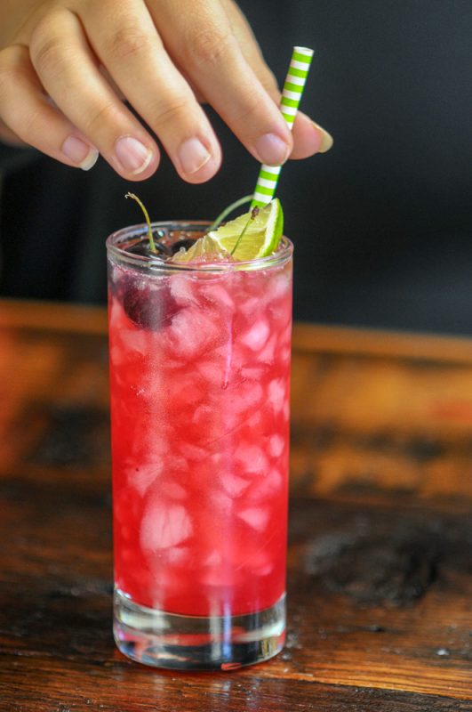 Cherry Cocktail in a highball with cherries and a lime, with a hand on a green and white straw