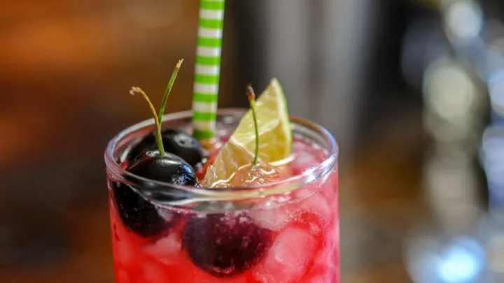 Bright pink cocktail in highball glass, cherries, lime wedge and green/white striped straw