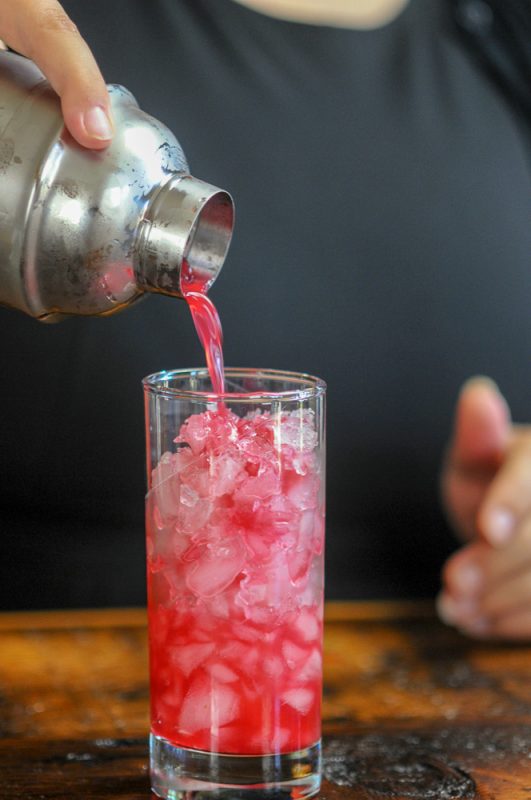 Ginger Cherry Smash Cocktail, pink cocktail being poured from shaker into highball glass filled wiht ice
