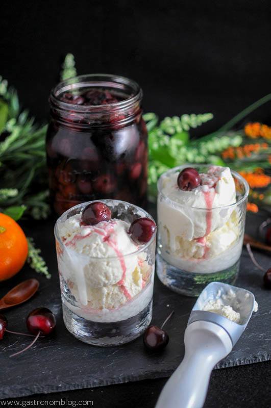 No Churn Old Fashioned Ice Cream in rock glasses with brandied cherries on top and in a jar. Orange, wooden spoons and cherries on slate board