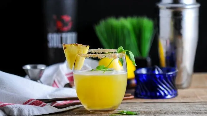 Pina de Fuego Cocktail, yellow cocktail with pineapple slice, blue juicer, shaker, grass and tequila bottle in background
