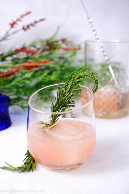 Rosada Cocktail with rosemary sprig and ice cube. Mixing glass and bar spoon with flowers in background