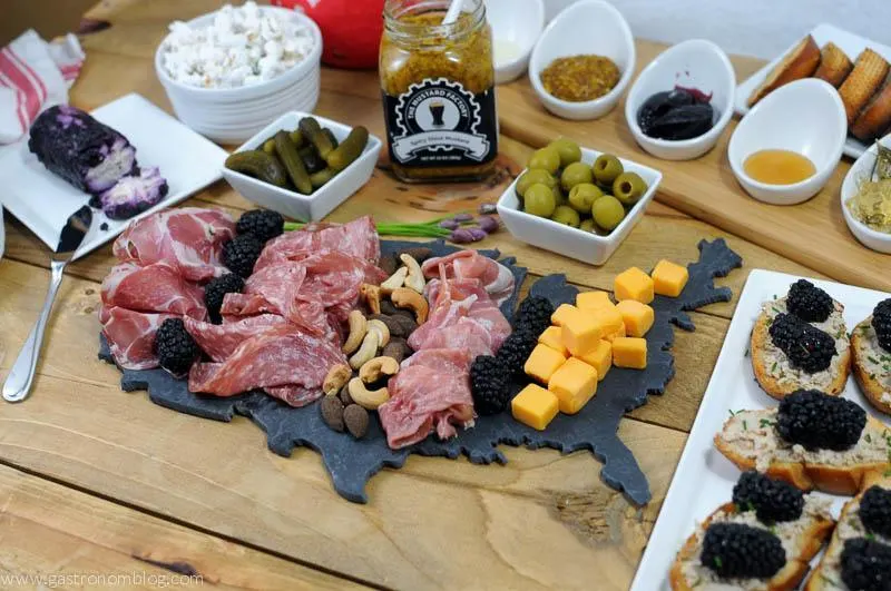 Charcuterie spread on a USA shaped slate board. Cheese, pickles, olives, crostini and spreads in white ceramic containers in the background