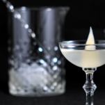 White cocktail in coupe with ginger slice, glass with ice and bar spoon in background
