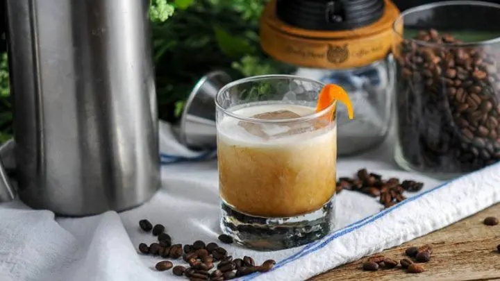 Cold Brew Bourbon Cocktail in rocks glass on white napkin with coffee beans. Coffee grinder and French press behind