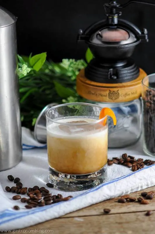 Cold brew coffee cocktail in a rocks glass with cream being poured into it. Coffee grinder, white napkin and coffee beans in the background