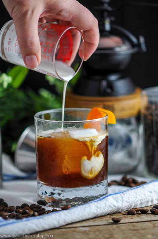 Cold brew coffee cocktail in a rocks glass with cream being poured into it. Coffee grinder, white napkin and coffee beans in the background