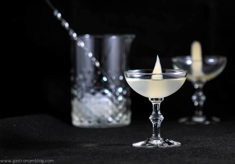 Sake cocktali in coupe with pickled ginger garnish. Mixing glass with bar spoon and another coupe in front of black background
