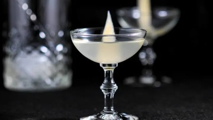 cocktail in coupe with slice of pickled ginger, another empty coupe in back with mixing glass and bar spoon in front of a black background