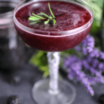 Purple cocktail in coupe, lavender greens garnish, flowers behind