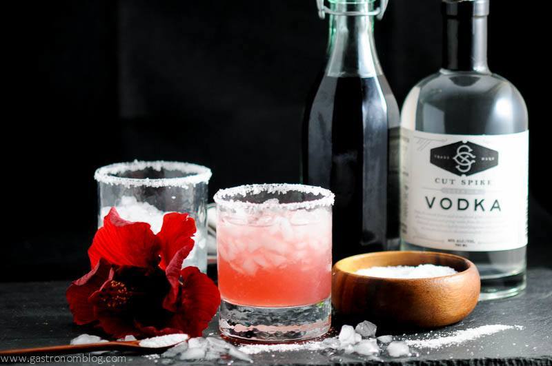 Hibiscus Salty Dog in rocks glasses with hibiscus simple syrup and vodka bottles in background with Hibiscus flower