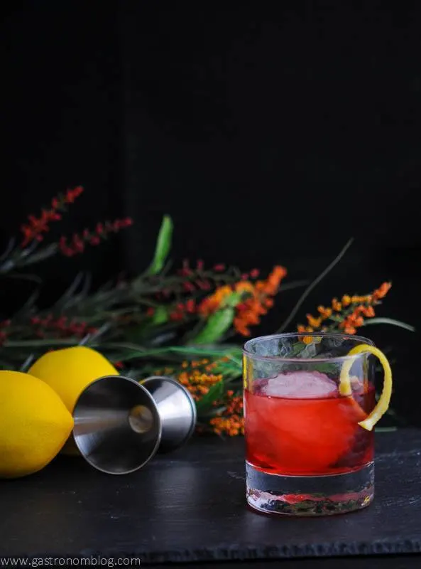 The Boulivardier -red cocktail in rocks glass with ice ball and lemon peel. Flowers, lemon and jigger in background