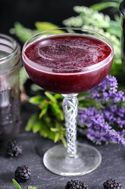 Brandy and Blackberry Lavender Shrub Cocktail -purple cocktail in coupe. Blackberries and blackberry shrub in jar, purple and white flowers in the back