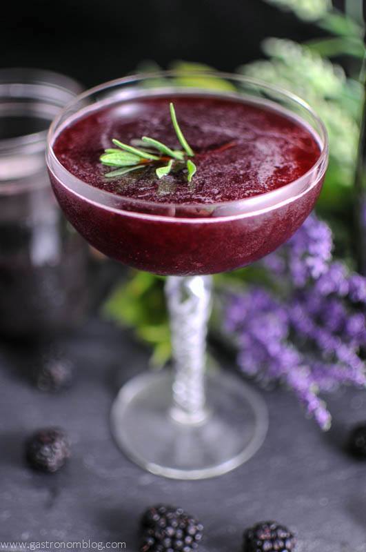 Purple cocktail in coupe with lavender leaves. Flowers behind coupe