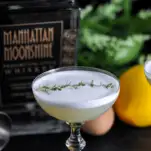 White foam topped cocktail in coupe, thyme on top, whiskey bottle, lemon and greenery in background