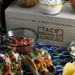 Tacos in a taco holder, box and bowls behind