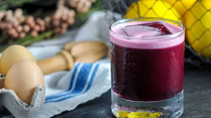 The Aronia Sour, maroon cocktail in rocks glass with square ice ball. Eggs, napkin, lemons and flowers in background
