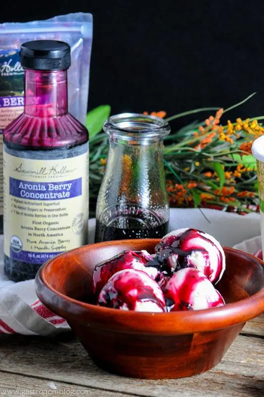 Aronia Berry Syrup on ice cream in a wooden bowl. Aronia berry bottle and bag in back with flowers