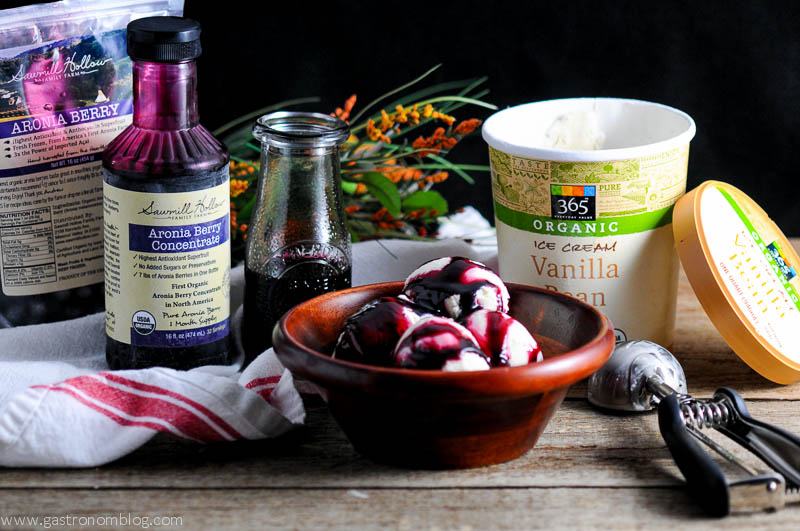 Aronia Berry Syrup on ice cream in wooden bowl. Aronia berry bottle and bag in background with ice cream container