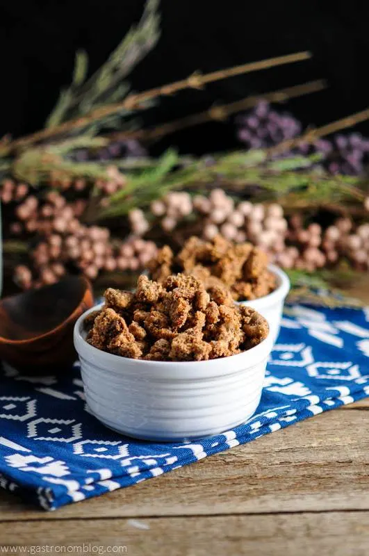 Bourbon Candied Pecans, these roasted nuts in white ceramic bowl on blue and white napkin, wood spoon and flowers in background