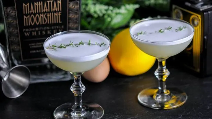 Thyme to Shine Cocktail in 2 coupes with white foam and thyme sprig on top. Bottle behind, lemon, flowers in background