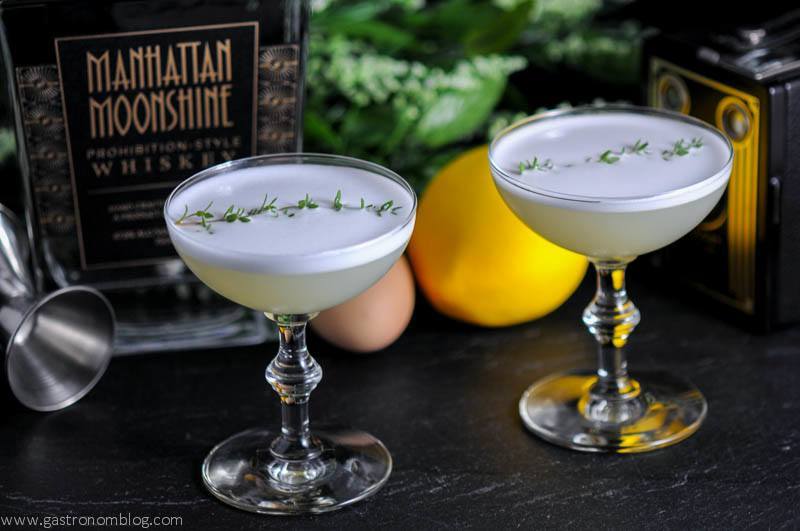 Thyme to Shine Cocktail in two cocktail coupes with egg, jigger, lemon and old camera in background with whiksey bottle