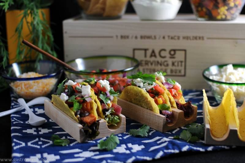 Sweet potato, black bean and corn tacos in wooden stands, glass bowls with ingredients behind. Wooden taco box and blue and white napkin