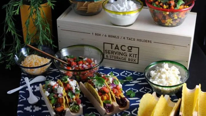 Sweet potato, black bean and corn tacos in wooden holders, with glass bowls of ingredeints behind. Taco box kit behind