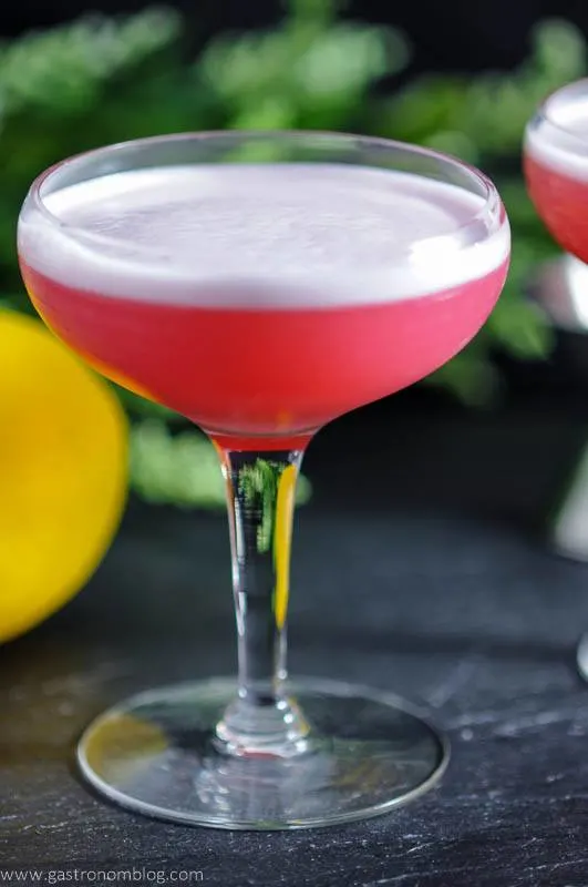 The Clover Club Cocktail - A gin and raspberry cocktail in a coupe with lemon and flowers in background