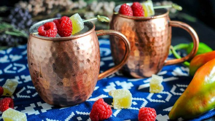 Hatch Chille and Raspberry Moscow Mule in copper mugs with raspberries and candied ginger. Raspberries and candied ginger with hatch chiles on blue and white napkin