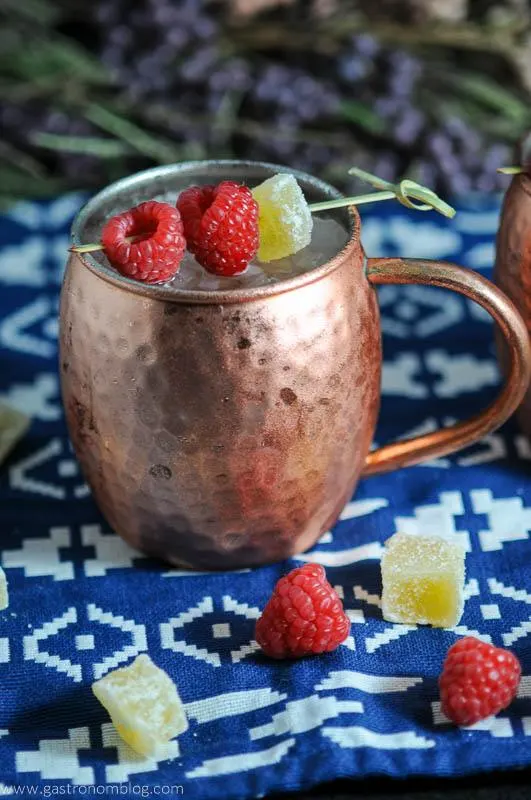Hatch Chille and Raspberry Moscow Mule in copper mug with raspberries and candied ginger on blue and white napkin