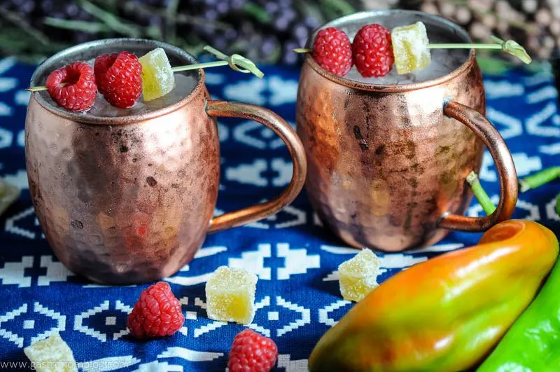 Hatch Chille and Raspberry Moscow Mule in copper mug with crystallized ginger and raspberries on blue and white napkin