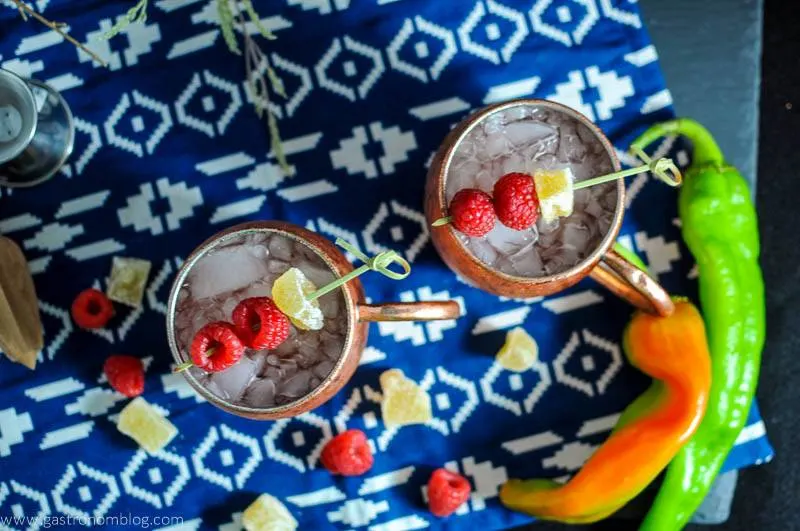 Hatch Chille and Raspberry Moscow Mule in copper mug with crystallized ginger and raspberries on blue and white napkin