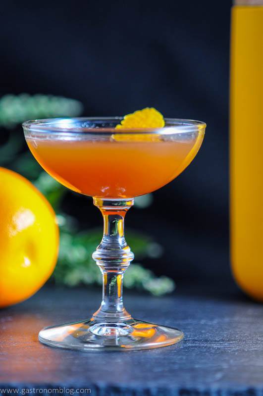 The Lucky Lucano cocktail, orange cocktail in coupe with orange peel, orange, flowers and yellow jar behind