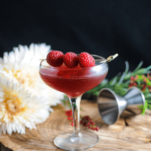 Pink cocktail in coupe with raspberries, on wood with flowers behind