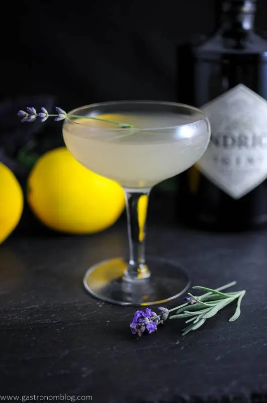 Lavender Bee's Knees Cocktail in coupe with lavender sprig. Lemon and flowers in background with gin bottle