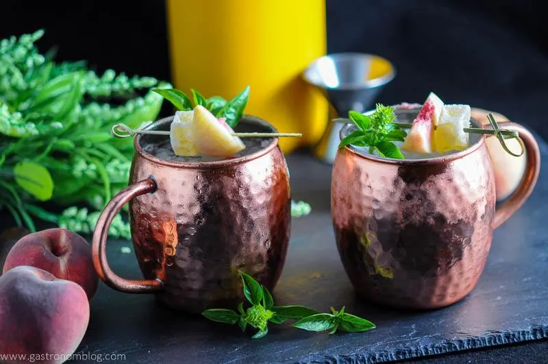 Peach and Basil Moscow Mule in copper mugs. Peaches, flowers, yellow container and jigger in background