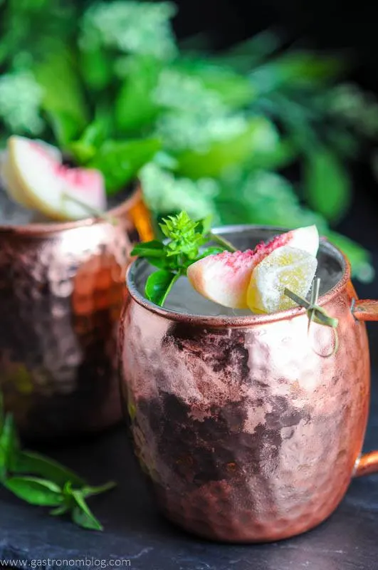 Peach and Basil Moscow Mule in copper mugs with peaches and basil on cocktail picks. Flowers in background