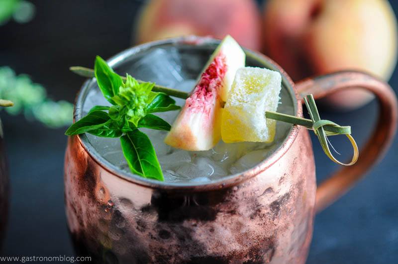 Peach and Basil Moscow Mule in copper mug with peach and candied ginger on cocktail pick with basil sprig