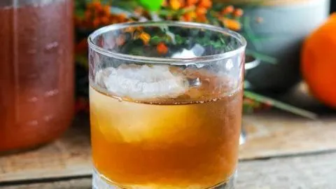 Peach Basil Bourbon Shrub Cocktail in a rocks glass with ice cube, shrub in jar behind, gray jar and orange flowers