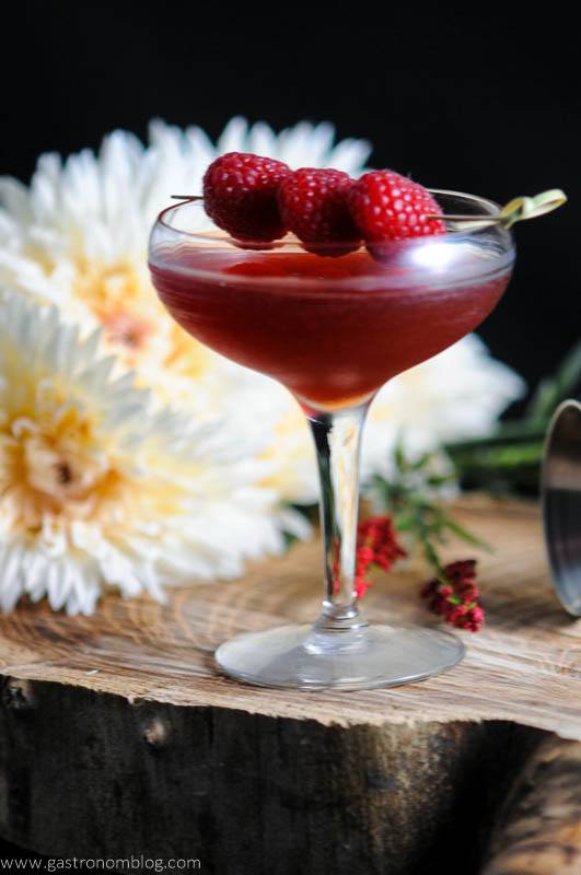 Royal Raspberry pink cocktail in coupe with rapsberries on pick. White flowers in backgorund