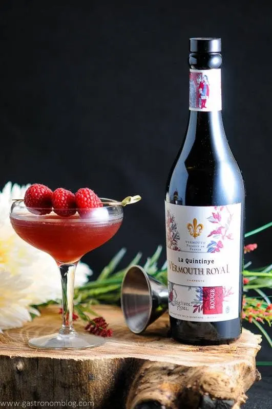 Red cocktail in coupe with raspberries on cocktail pick, vermouth bottle, jigger and flowers on wood platter