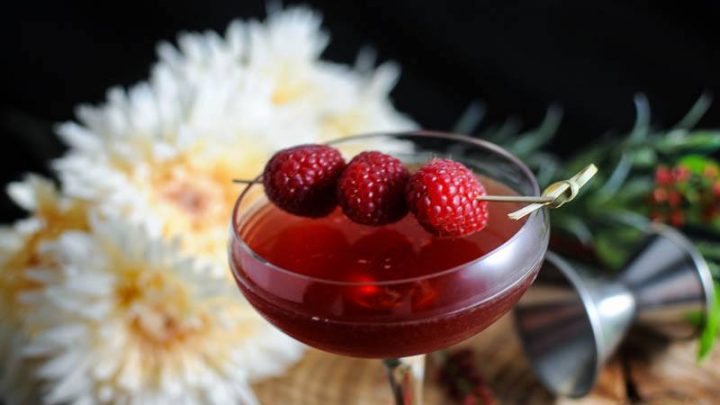 Royal Raspberry, pink cocktail in coupe with raspberries on cocktail pick. White flowers and jigger behind