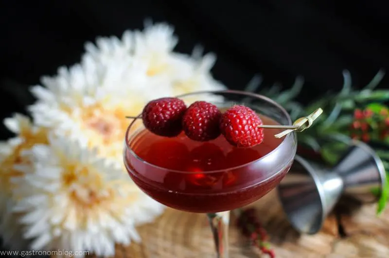 Raspberry Manhattan Cocktail in cocktail coupe. Flowers and jigger in background