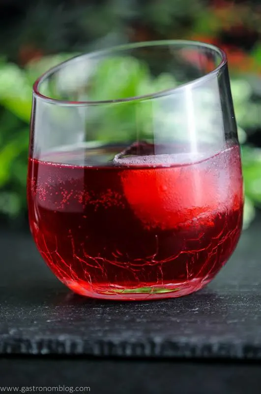 The Cranberry Sweet and Sour, red liquid in glass with large ice cube, flowers in background