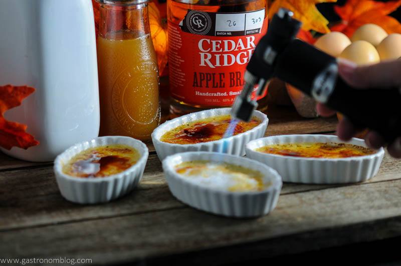 Apple Cider Brandy Creme Brulee in ceramic baking dishes with culinary torch flame. White bottle, jar of cider and apple brandy bottle in background