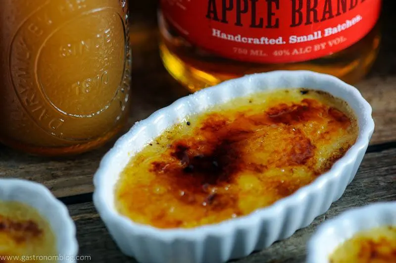 apple Creme brulee in white ramkeins, apples, fall leaves and brandy bottle behind