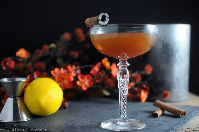 The Clove and Cider Cocktail in cocktail coupe. Metal container, cinnamon sticks, lemon, flowers and jigger in background.