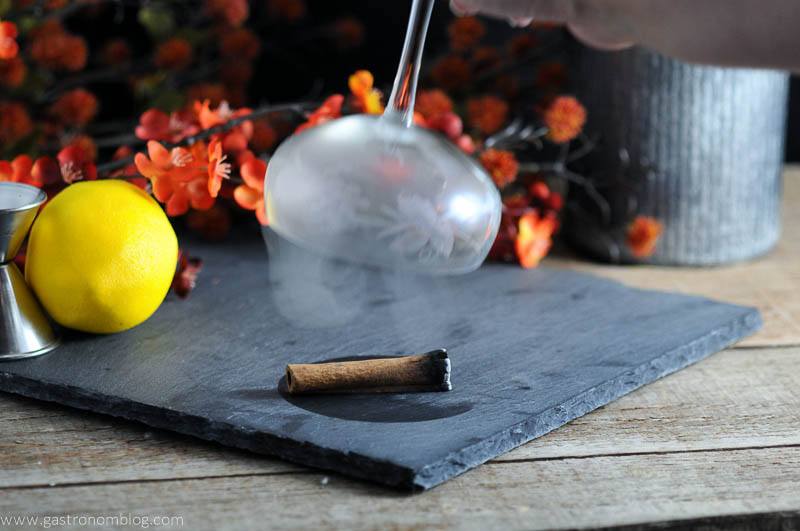 The Clove and Cider Cocktail glass being smoked with burning cinnamon. Flowers, lemon, and jigger in background