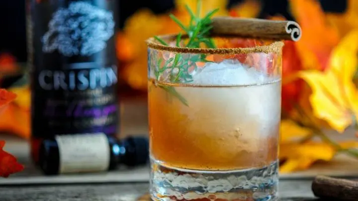 The Autumn Pear, brown cocktail in rocks glass with ice ball, rosemary sprig and cinnamon sticks. Cider bottle and bitters with autumn leaves in back